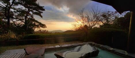 A hot spring area with a wooden deck, a stone hot tub, and a backdrop of trees and hills.