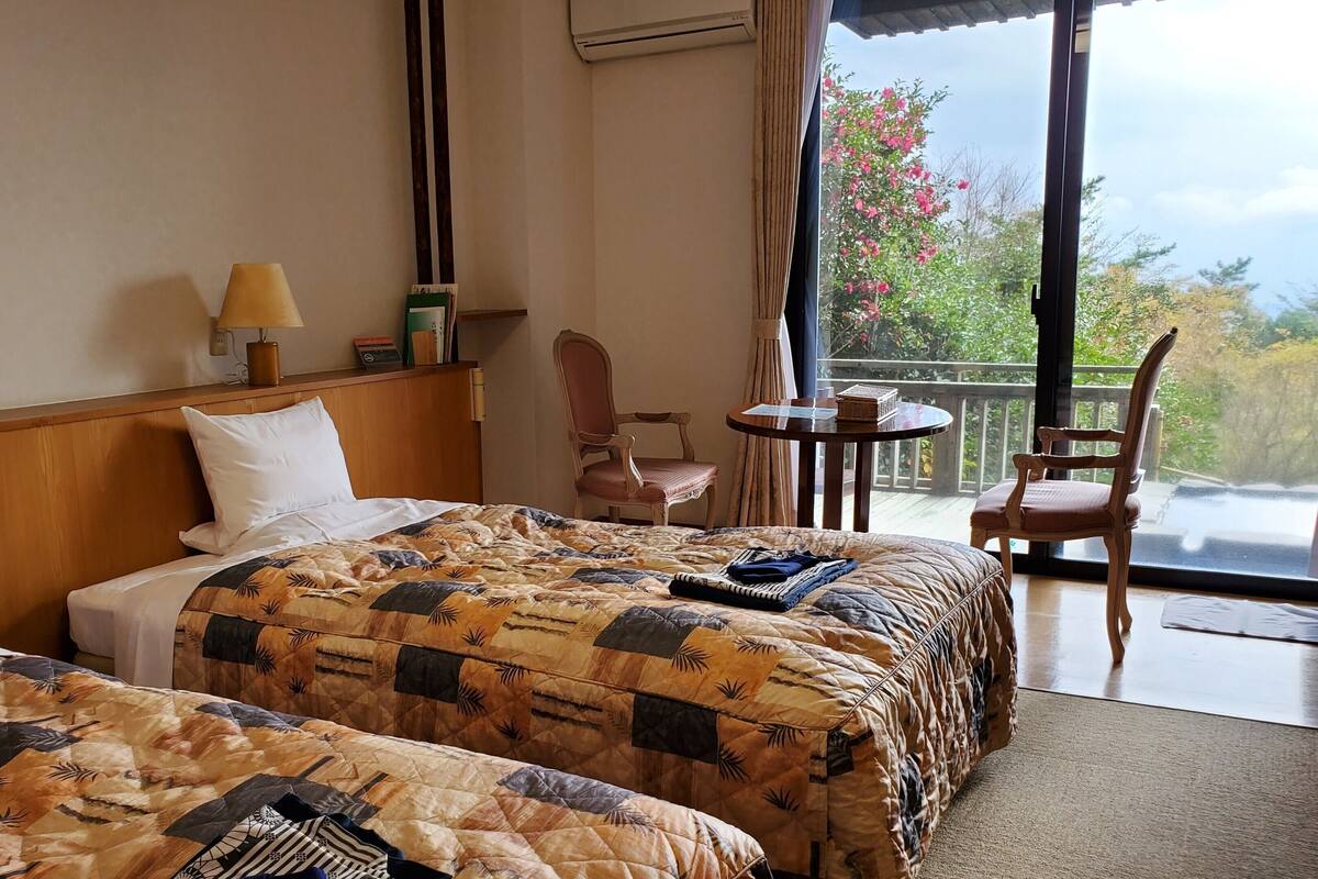 Une chambre d’hôtel avec deux lits, une tête de lit en bois, une petite table avec une lampe, une chaise et un balcon donnant sur des arbres et des fleurs.