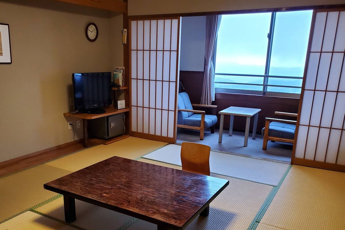 Une pièce de style japonais traditionnel, avec un sol recouvert de tatamis, une télévision, une horloge et une vue sur l’extérieur à travers des panneaux coulissants.