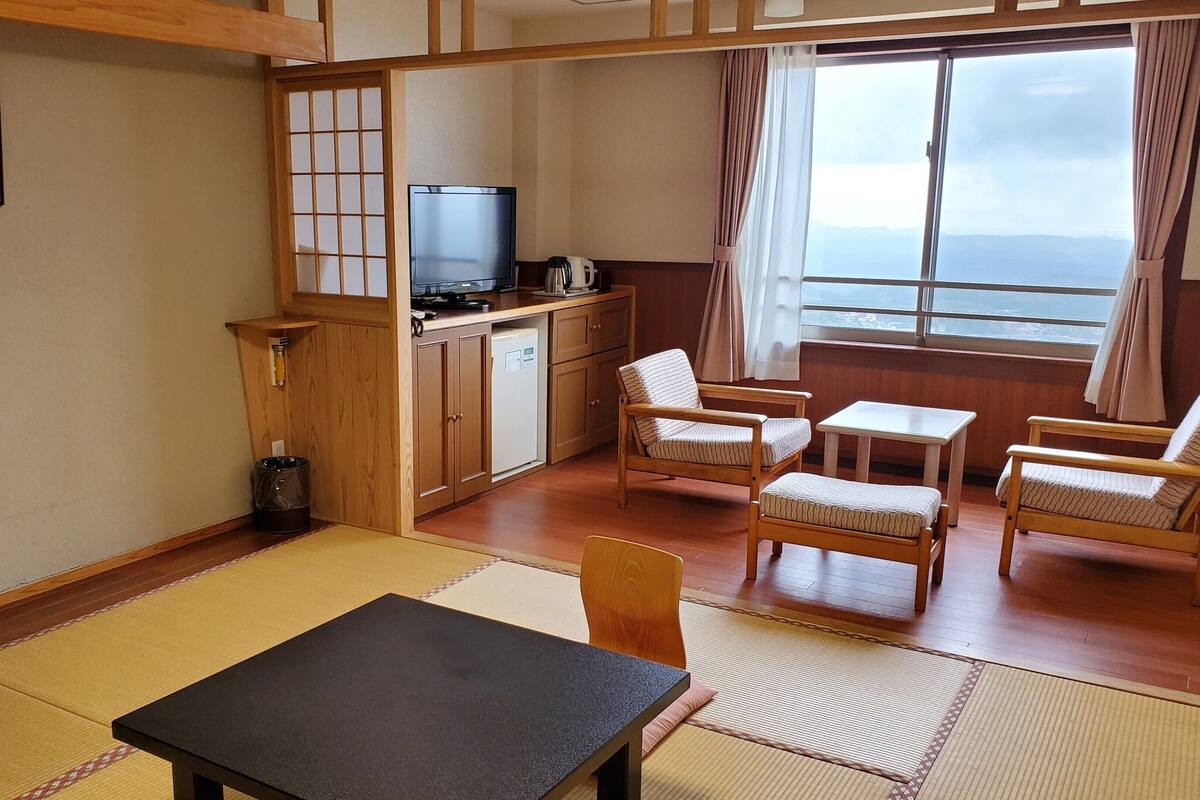 Une pièce avec un sol recouvert de tatamis, une table noire, des chaises en bois, un téléviseur, une machine à café et une vue sur la mer par la fenêtre.