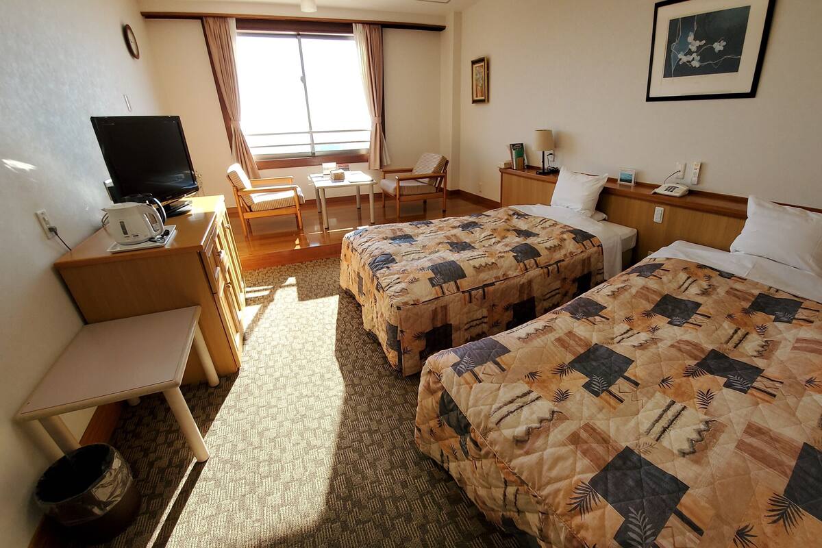 Une chambre d’hôtel avec deux lits, un bureau, une chaise, une télévision et une fenêtre avec des rideaux.