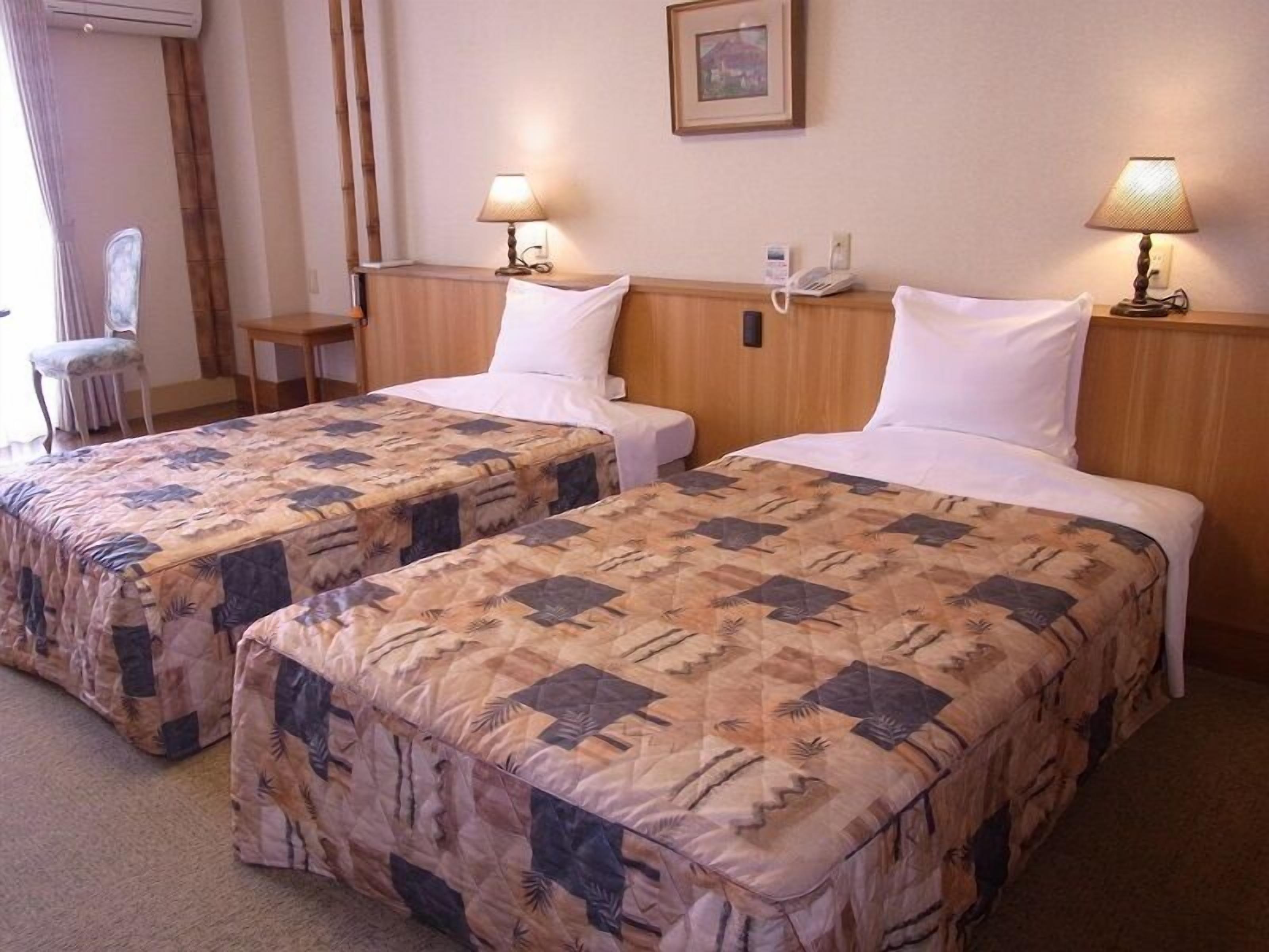 A hotel room with two beds, each with a patterned bedspread, bedside lamps, a telephone, and a framed picture on the wall.