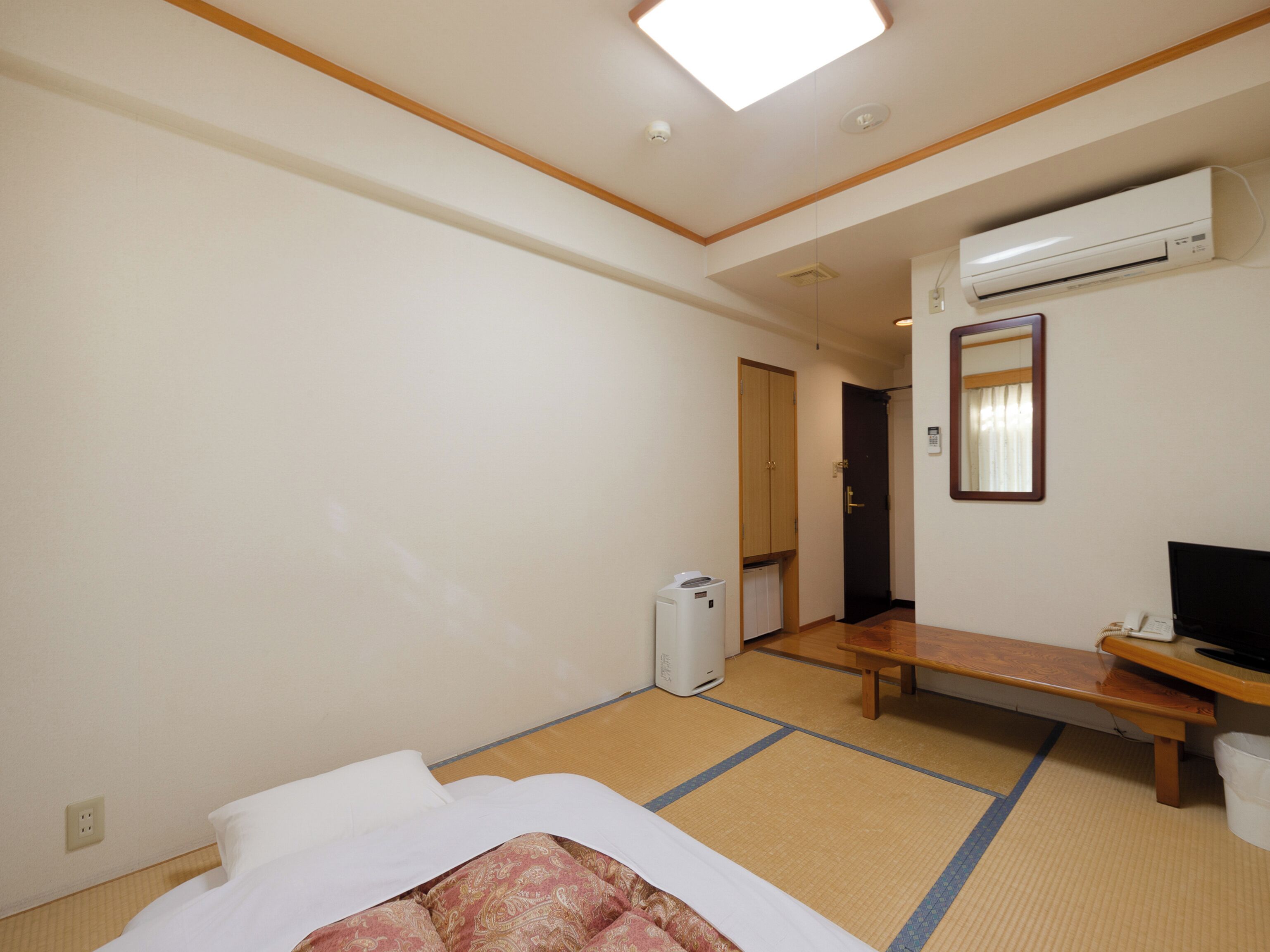A hotel room with a bed, a desk, an air conditioner, a mirror, and a door leading to another room.