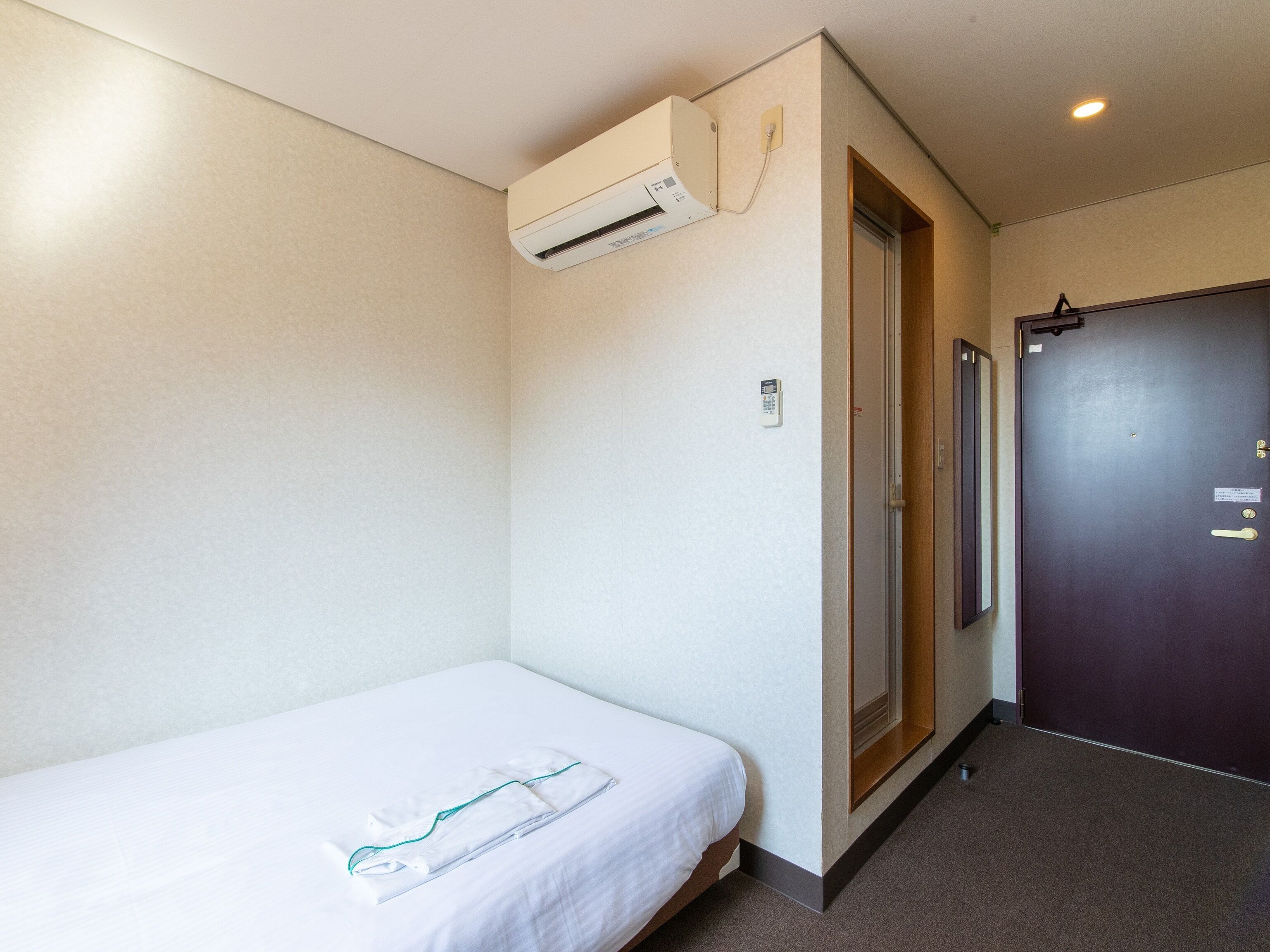 Una habitación pequeña y limpia con una cama individual, aire acondicionado de pared, una puerta con cerradura y un ventanal.