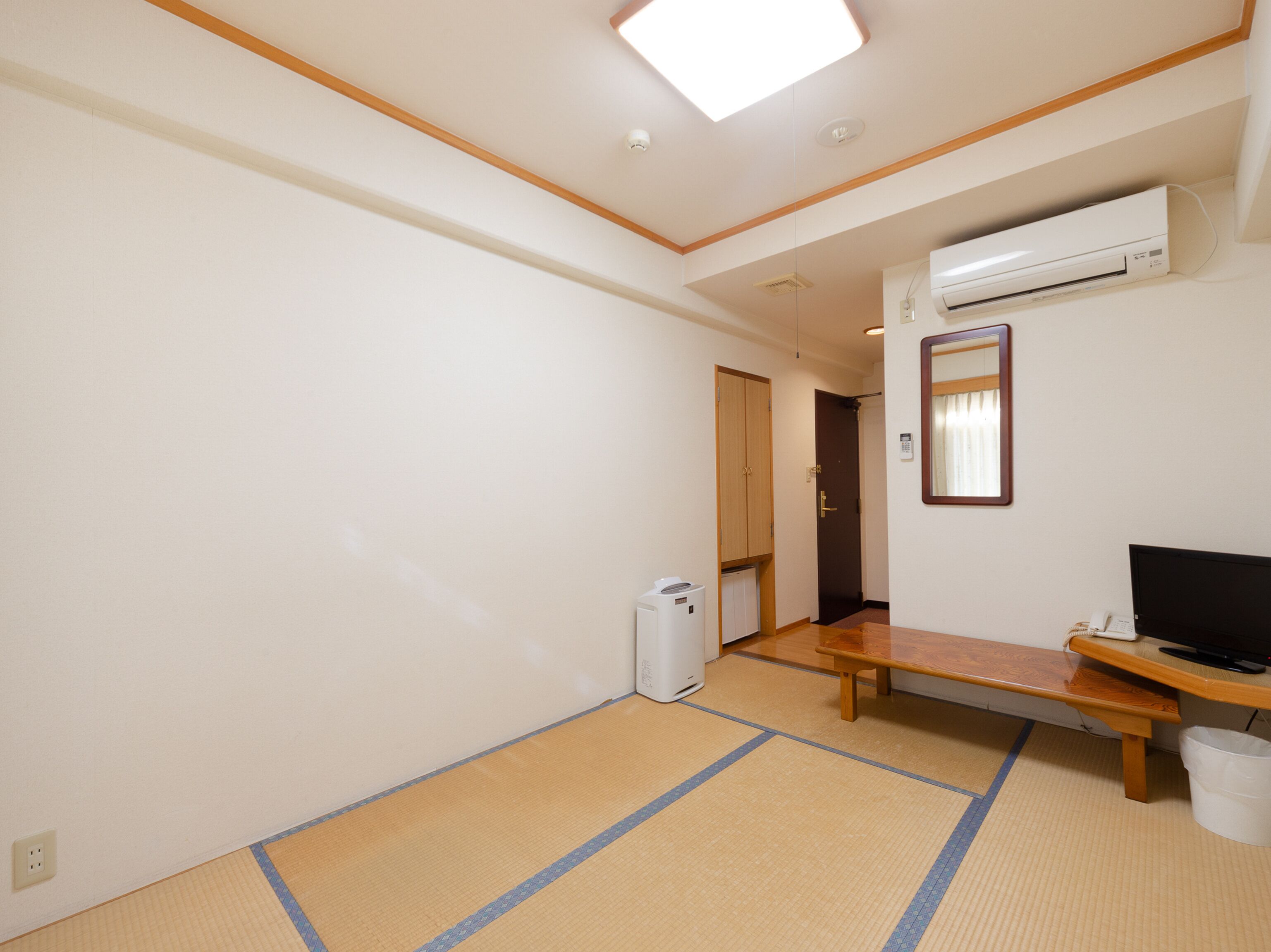 Ein Zimmer mit Tatami-Boden, einer Holzbank, einem Fernseher, einer Klimaanlage und einem kleinen Mülleimer.