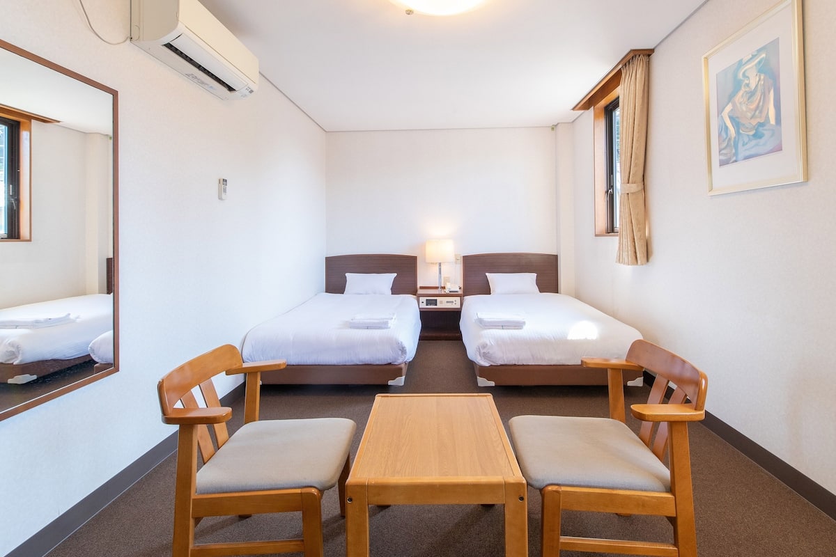 Une chambre d’hôtel avec deux lits simples, une petite table en bois, deux chaises, une table de chevet avec une lampe, un tableau au mur et un appareil de climatisation.
