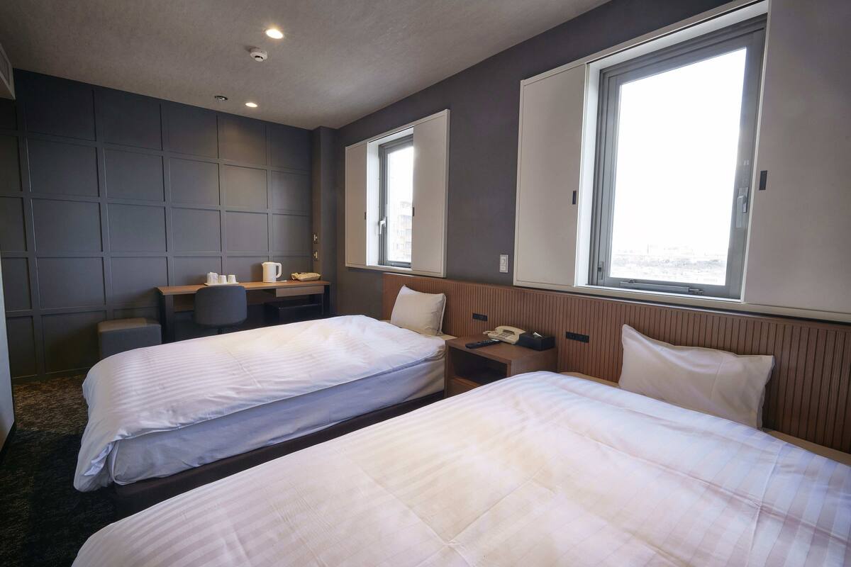 A hotel room with two beds, a desk, a chair, and a window.