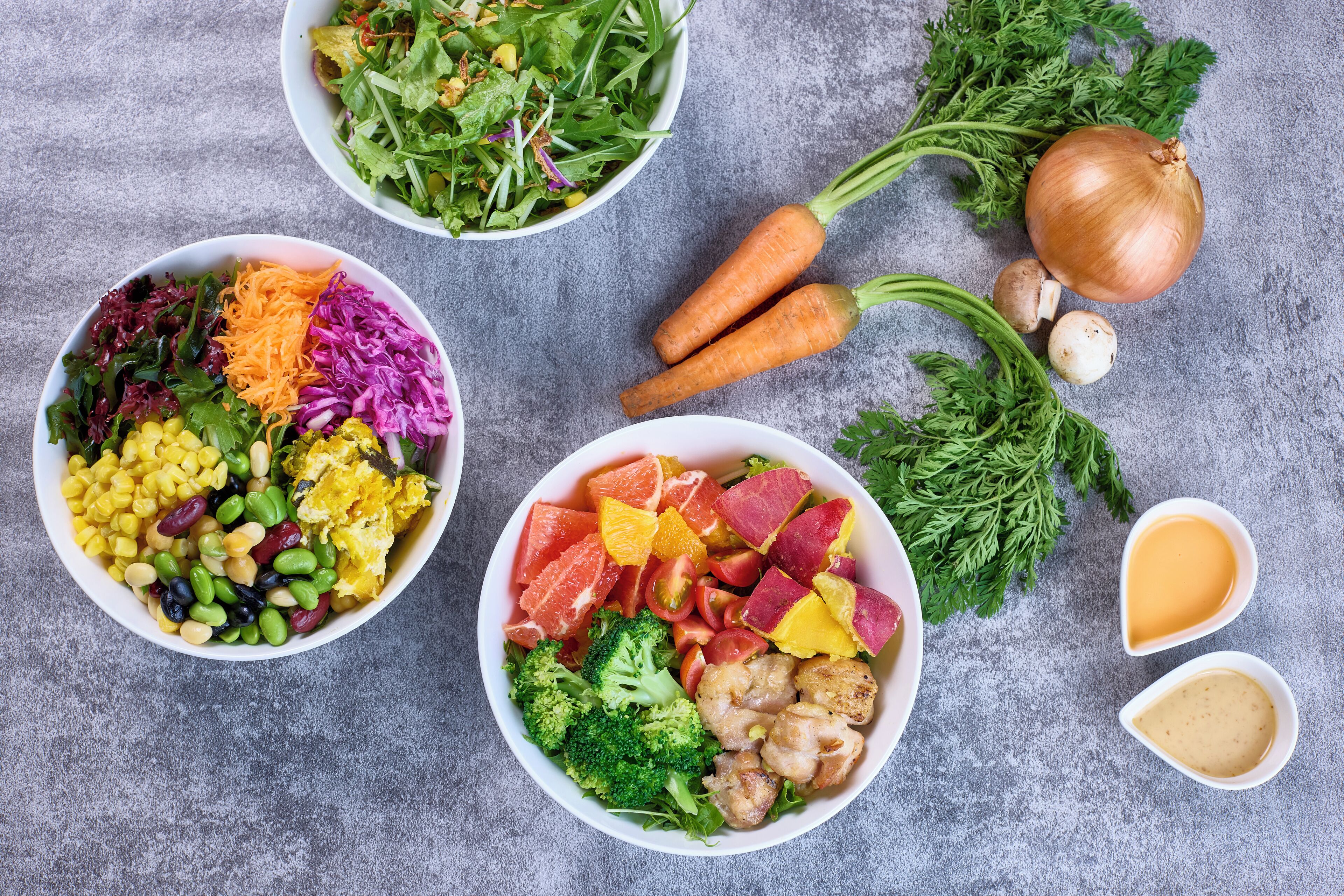 Two bowls of colorful salads with various vegetables and fruits, a whole onion, carrots, and parsley on a gray surface.