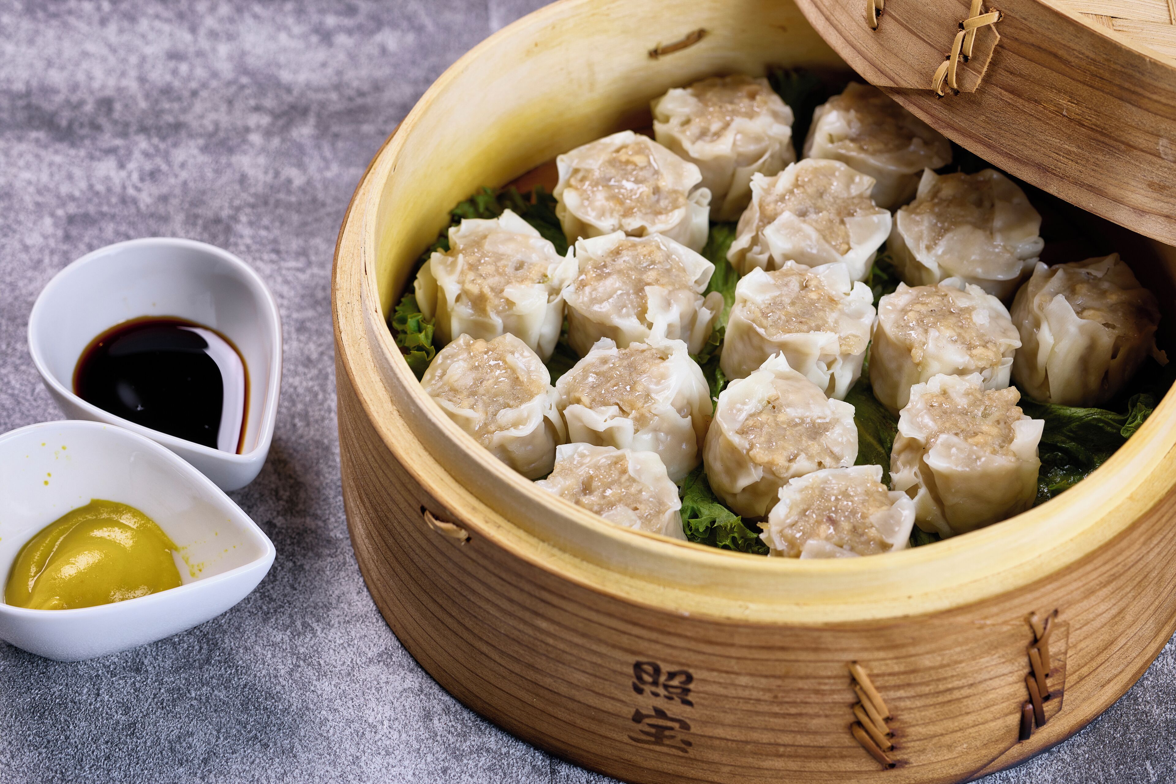 Um cestinho de bambu cheio de dumplings, uma pequena tigela de molho de soja e uma pequena tigela de mostarda.
