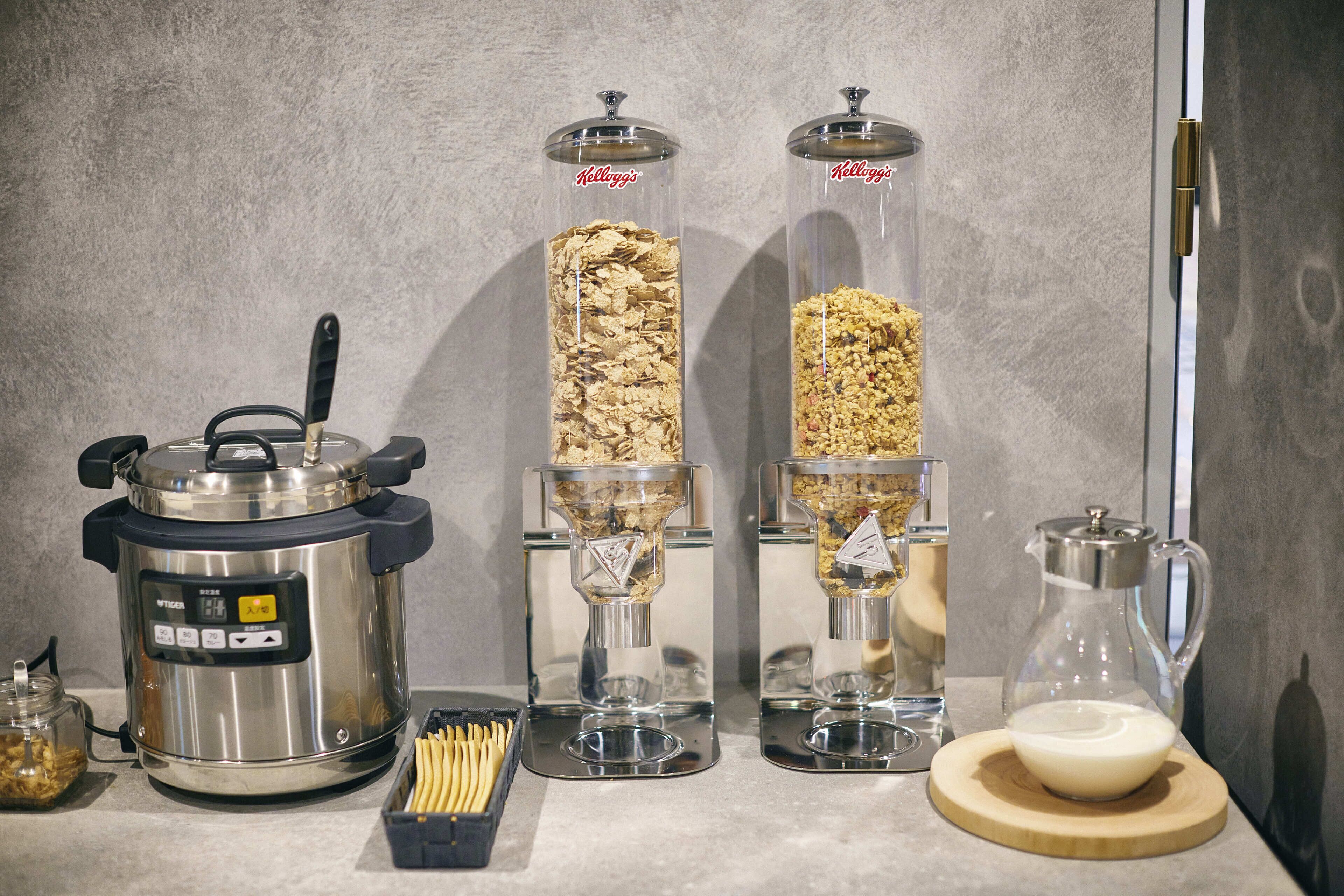 A kitchen area with a rice cooker, two glass containers with lids, a coffee maker, and a bowl of milk on a wooden board.