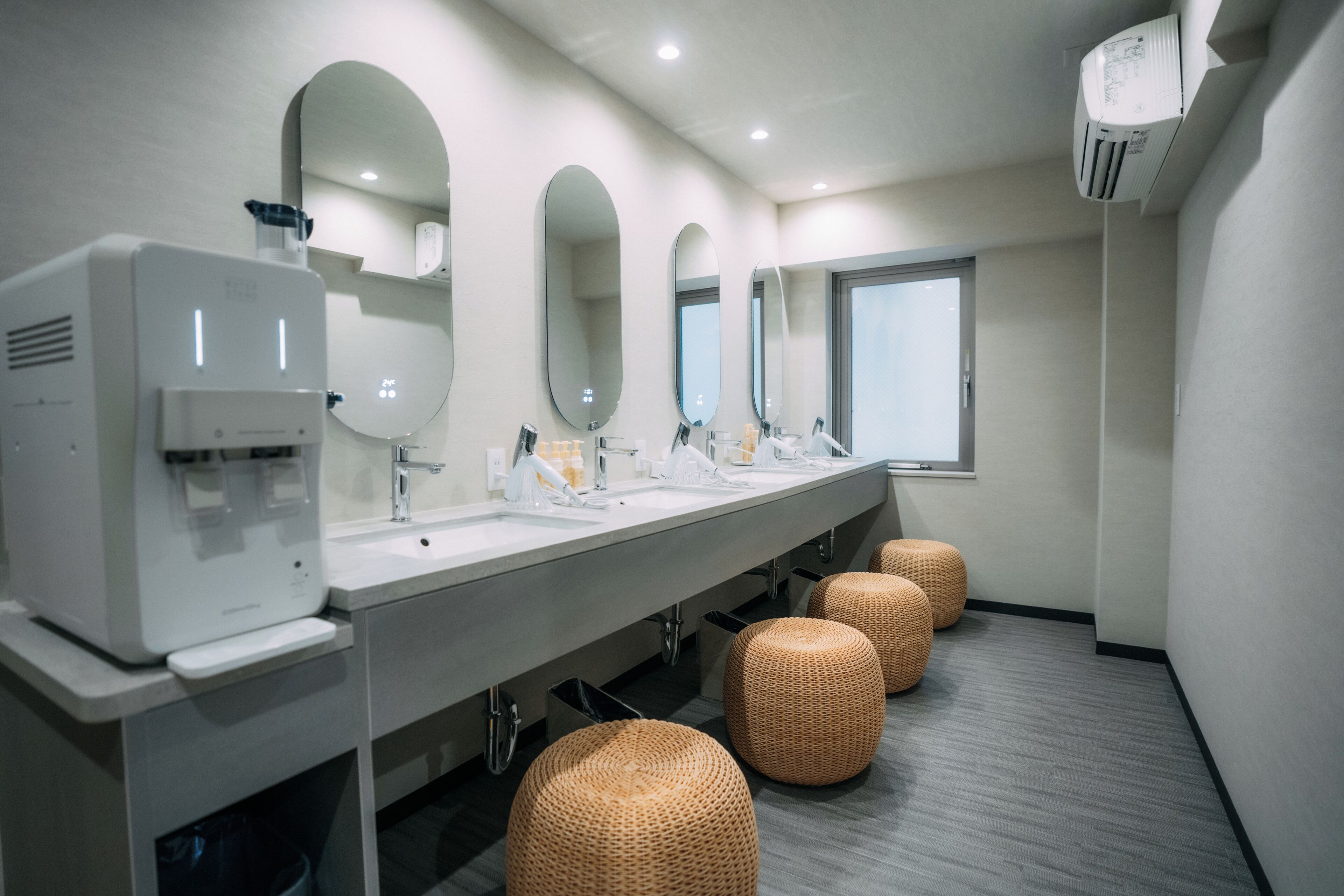 A modern bathroom with three sinks, a hand dryer, and three large mirrors. There are three woven stools and a window with blinds.