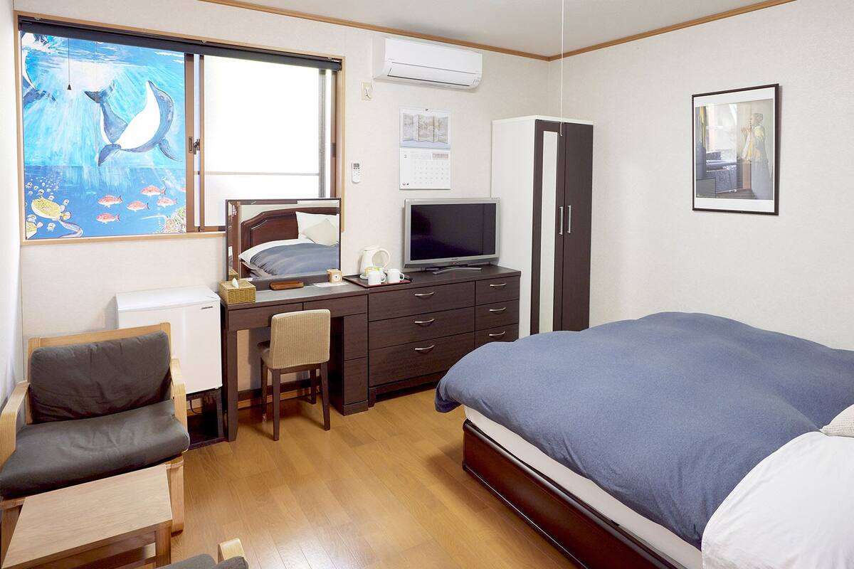 Une chambre à coucher comprenant un lit, un bureau avec une chaise, une télévision, une armoire et une fenêtre décorée d’œuvres d’art.
