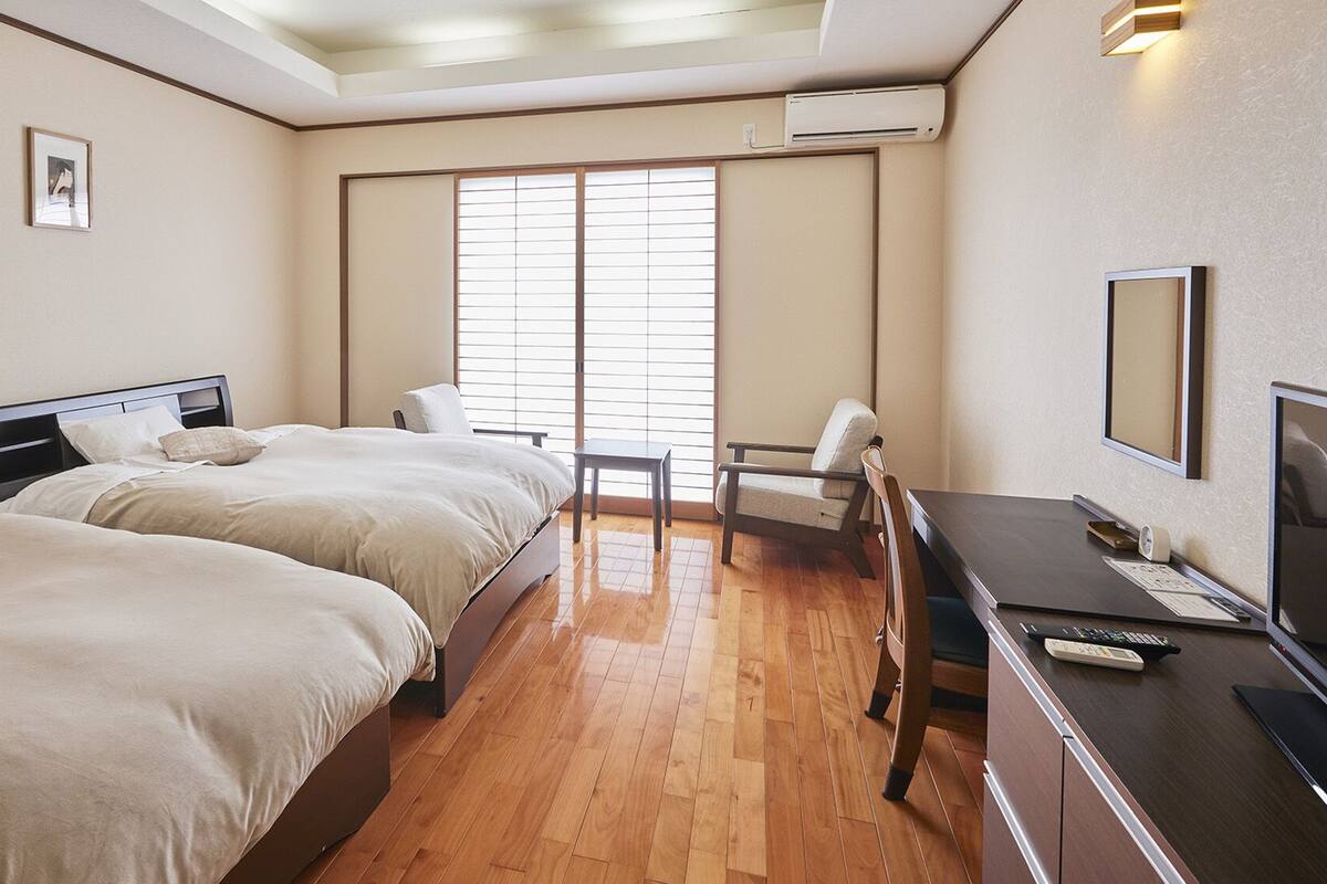 Une chambre d’hôtel avec deux lits, un bureau, une chaise, une télévision et une fenêtre avec des stores.