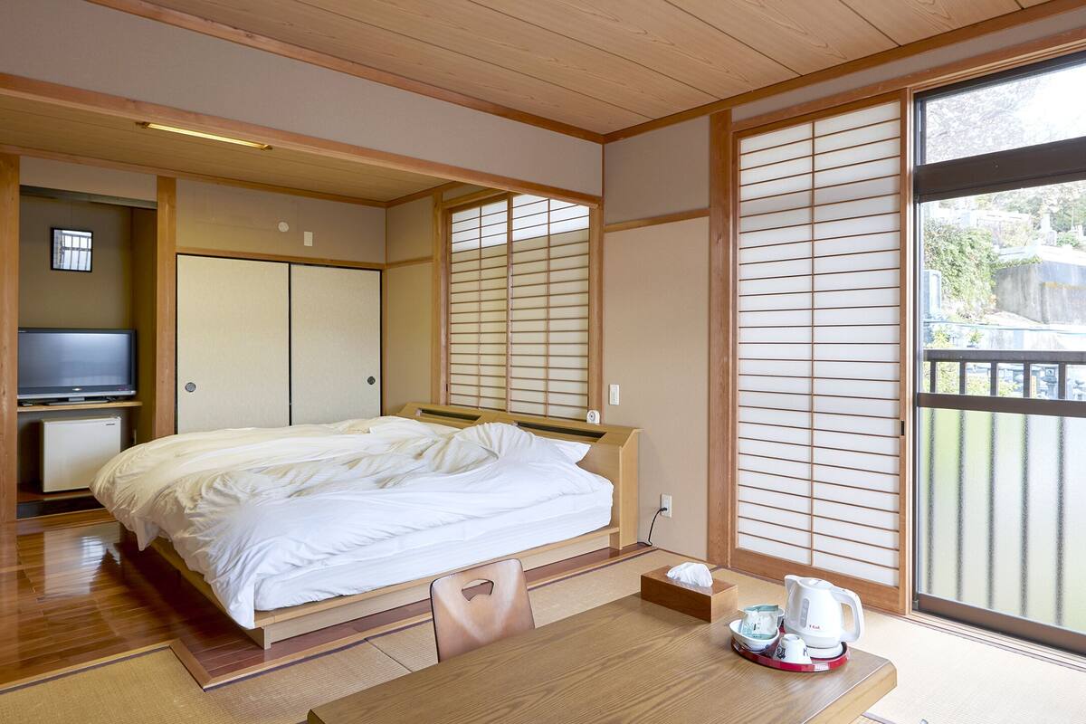 Une chambre de style japonais traditionnel, avec un sol recouvert de tatamis, un lit avec une literie blanche, une télévision, un petit réfrigérateur et une porte coulissante dotée de panneaux shoji.