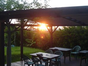 Eine Terrasse im Freien mit Tischen und Stühlen, einer Holz-Pergola und Blick auf den Sonnenuntergang.