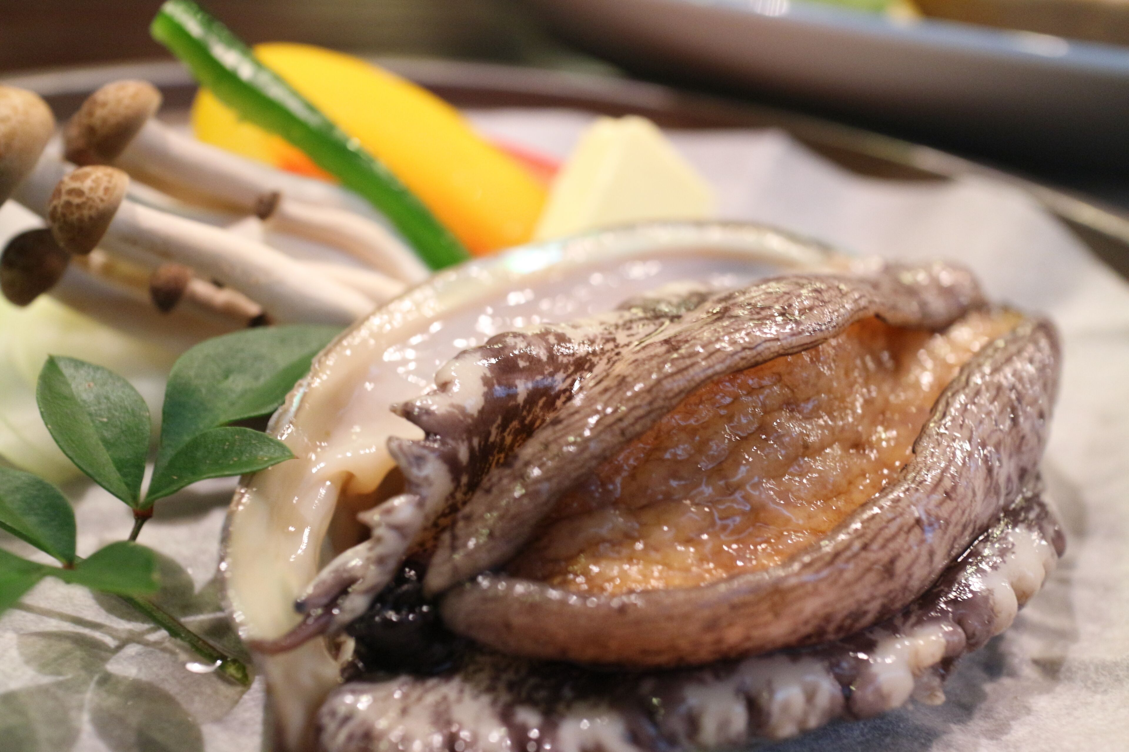 A close-up of a dish with a shell, possibly a seafood item, accompanied by some green leaves and a yellow object in the background.