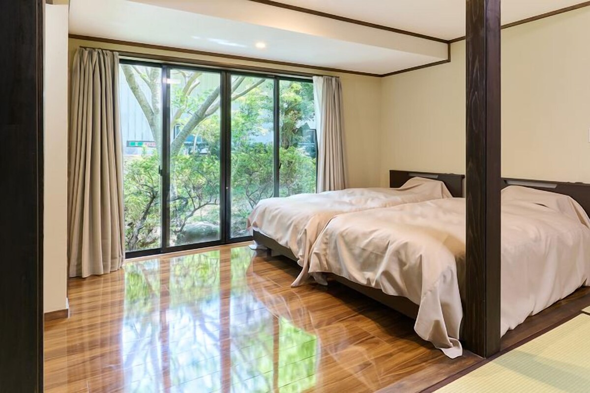 A bedroom with a large sliding glass door leading to a garden, a bed with a wooden headboard, and a tatami mat in the corner.