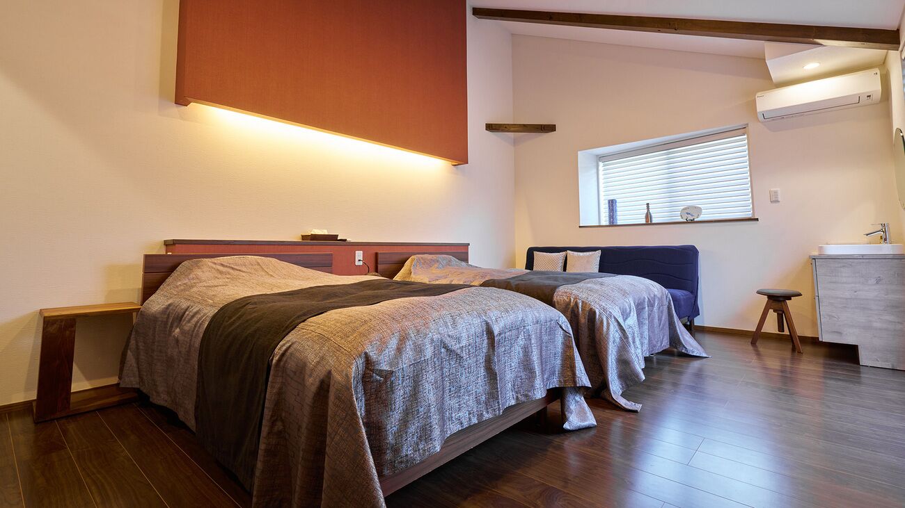 Un dormitorio con dos camas, una mesita de noche, un taburete pequeño, una ventana con persianas y un equipo de aire acondicionado.