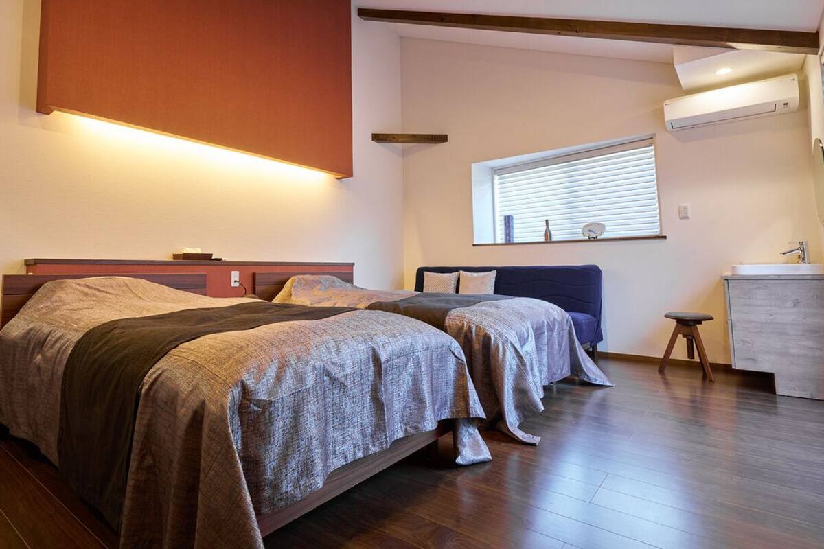 Un dormitorio con dos camas, una mesita de noche, un taburete pequeño, una ventana con persianas y un equipo de aire acondicionado.
