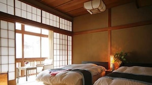 Um quarto japonês tradicional com tatames, duas camas, uma janela com portas deslizantes shoji e uma pequena mesa com uma cadeira.