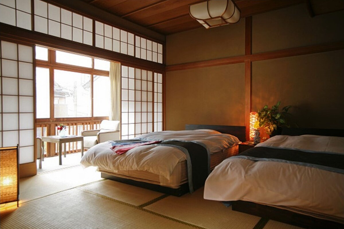 A traditional Japanese-style room with tatami flooring, a low wooden ceiling, and sliding shoji doors. There are two beds with white bedding, a small table with a chair, and a lamp on the nightstand.
