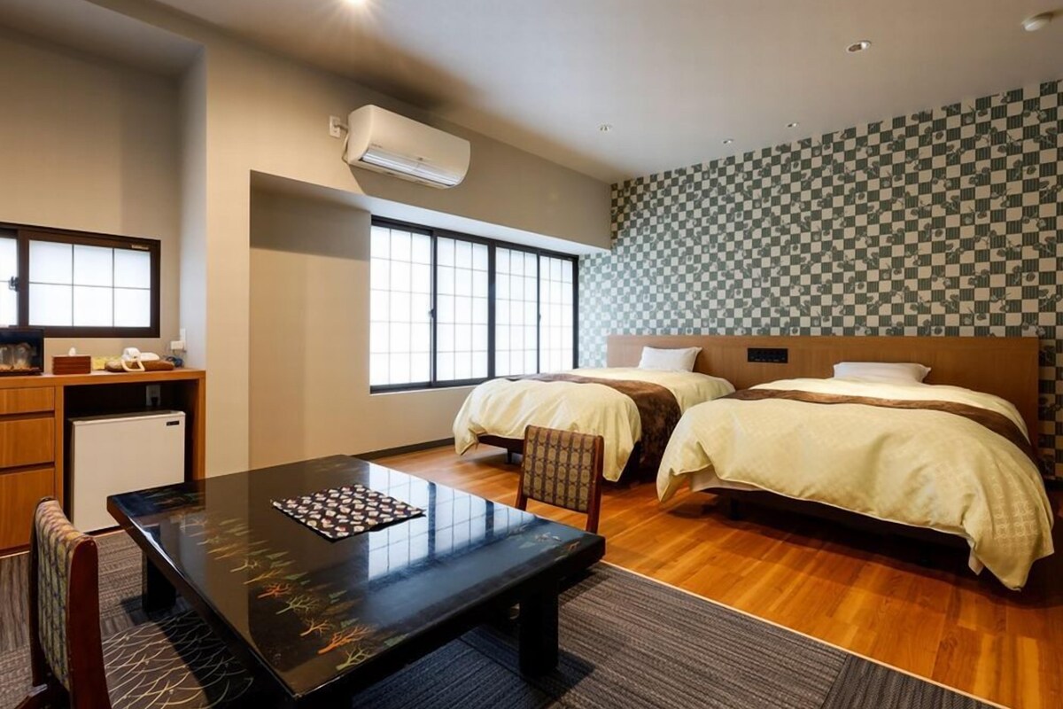 A hotel room with two beds, a dining table, a small kitchenette, and a large window.
