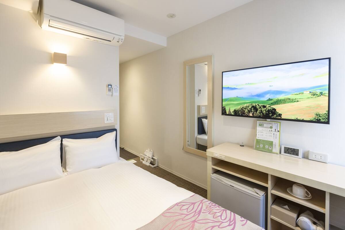 Ein Hotelzimmer mit einem Bett, einem an der Wand befestigten Fernseher, einem Schreibtisch mit Spiegel und einem kleinen Kühlschrank.