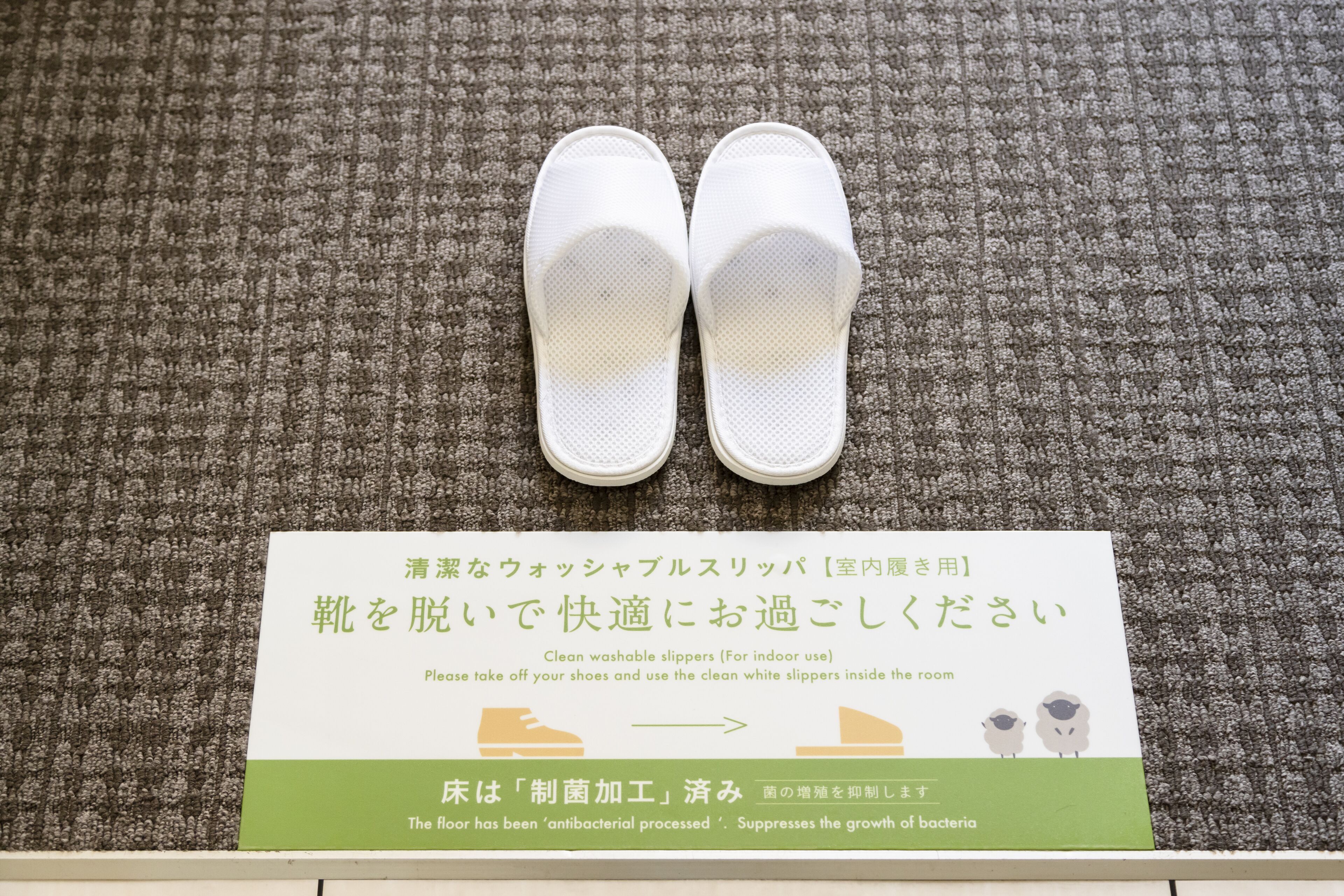 Ein Paar weiße Hausschuhe auf einem Teppichboden, mit einem Schild auf Japanisch, das zum Tragen in Innenräumen auffordert.