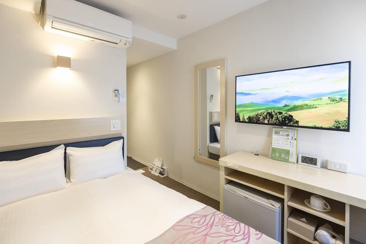 Ein Hotelzimmer mit Bett, Fernseher, der eine Landschaft zeigt, Spiegel und einer kleinen Küchenzeile.