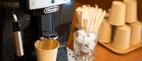 A coffee machine with a cup under the spout, a control panel, and a stack of disposable cups and a glass with a spoon on a counter.