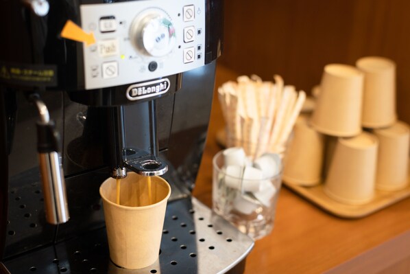 A coffee machine with a cup under the spout, a control panel, and a stack of disposable cups and a glass with a spoon on a counter. - Beppu Daiiti Hotel (Beppu)