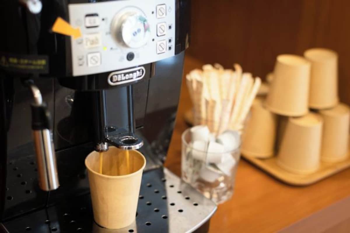 A coffee machine with a cup under the spout, a control panel, and a stack of disposable cups and stirrers on the table.