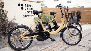 En gul cykel med en korg, parkerad framför en byggnad med en skylt där det står: ”BEPPU DAII HOTEL, ESTRABLERAT 1972”.