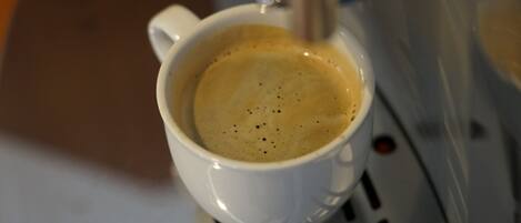Una taza blanca de café sobre una superficie metálica, con una máquina de café al fondo.