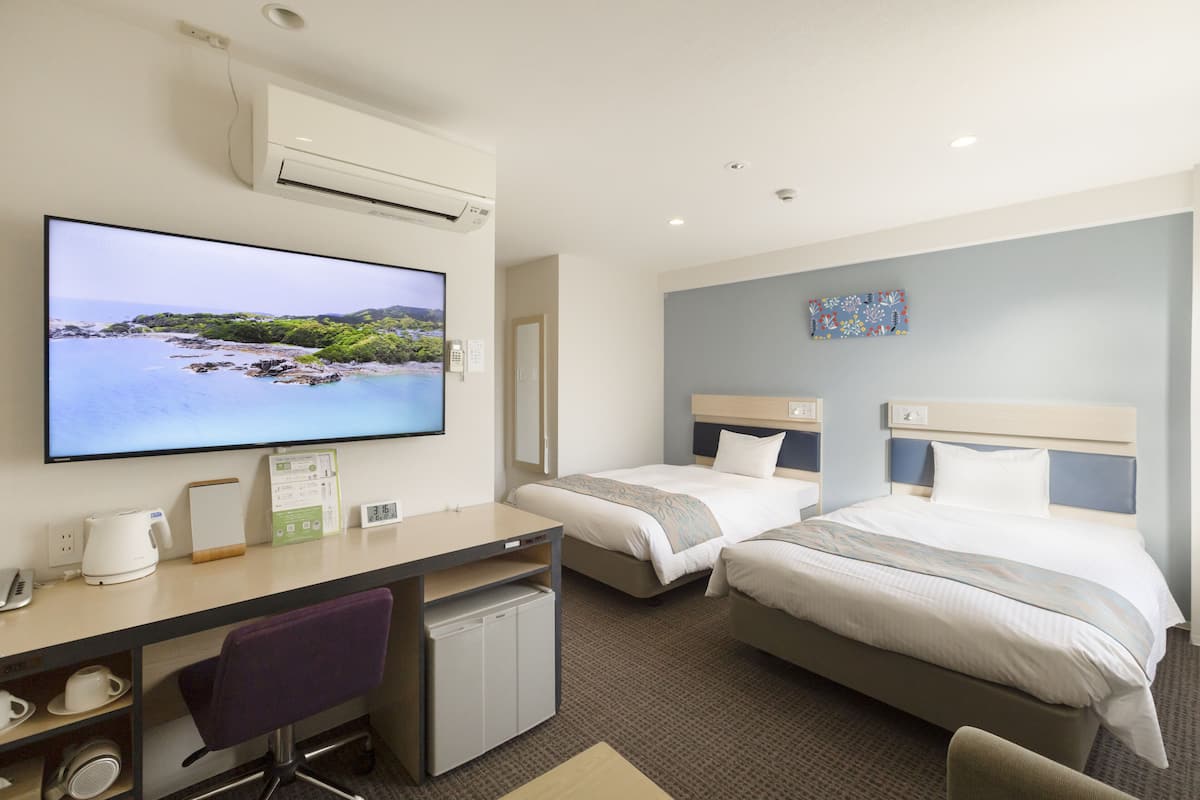 Ein Hotelzimmer mit Fernseher, Schreibtisch, zwei Betten, einer kleinen Kühlschrank und einem Sessel.