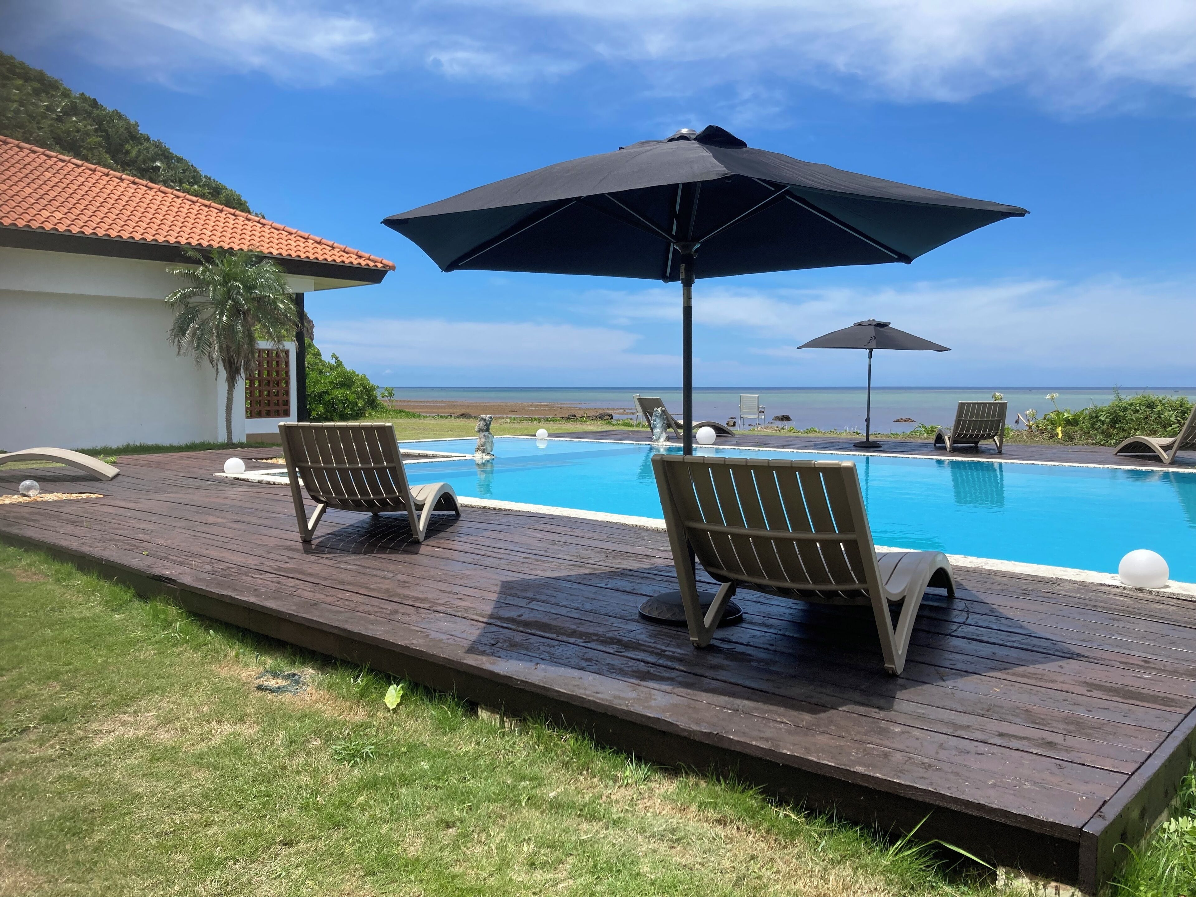 A wooden deck with two lounge chairs and a large umbrella, overlooking a swimming pool and the ocean.