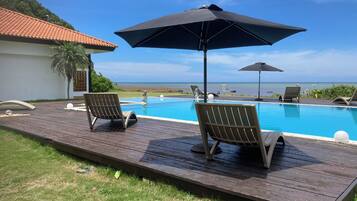 A wooden deck with two lounge chairs and a large umbrella, overlooking a swimming pool and the ocean.