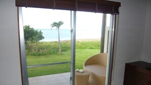A room with a sliding glass door leading to a balcony with a view of the ocean and a palm tree. There is a wicker chair and a small table on the balcony. - Eco Village Iriomote (Taketomi)