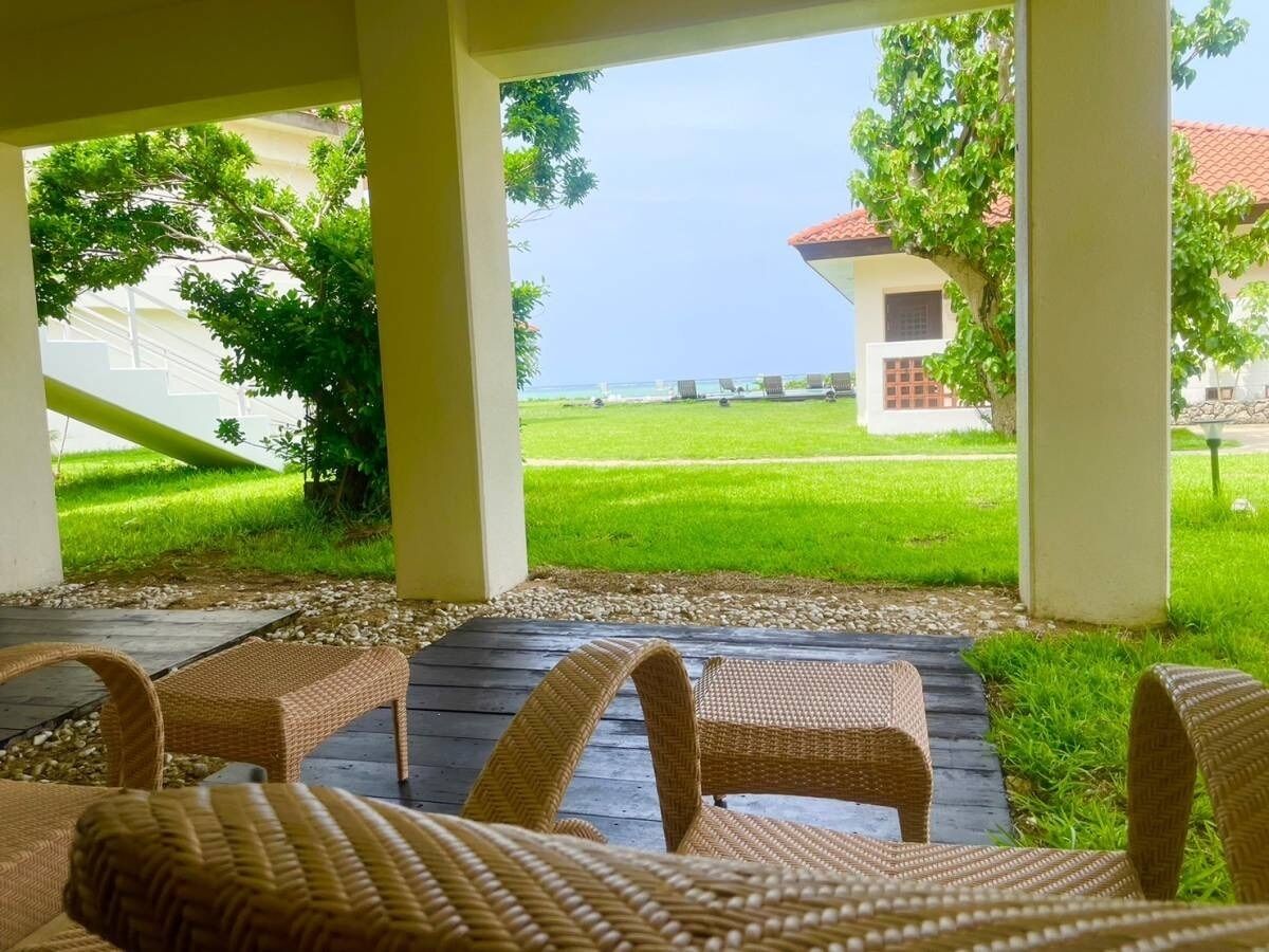 A covered outdoor seating area with wicker furniture, a view of a grassy lawn, and a building in the background.