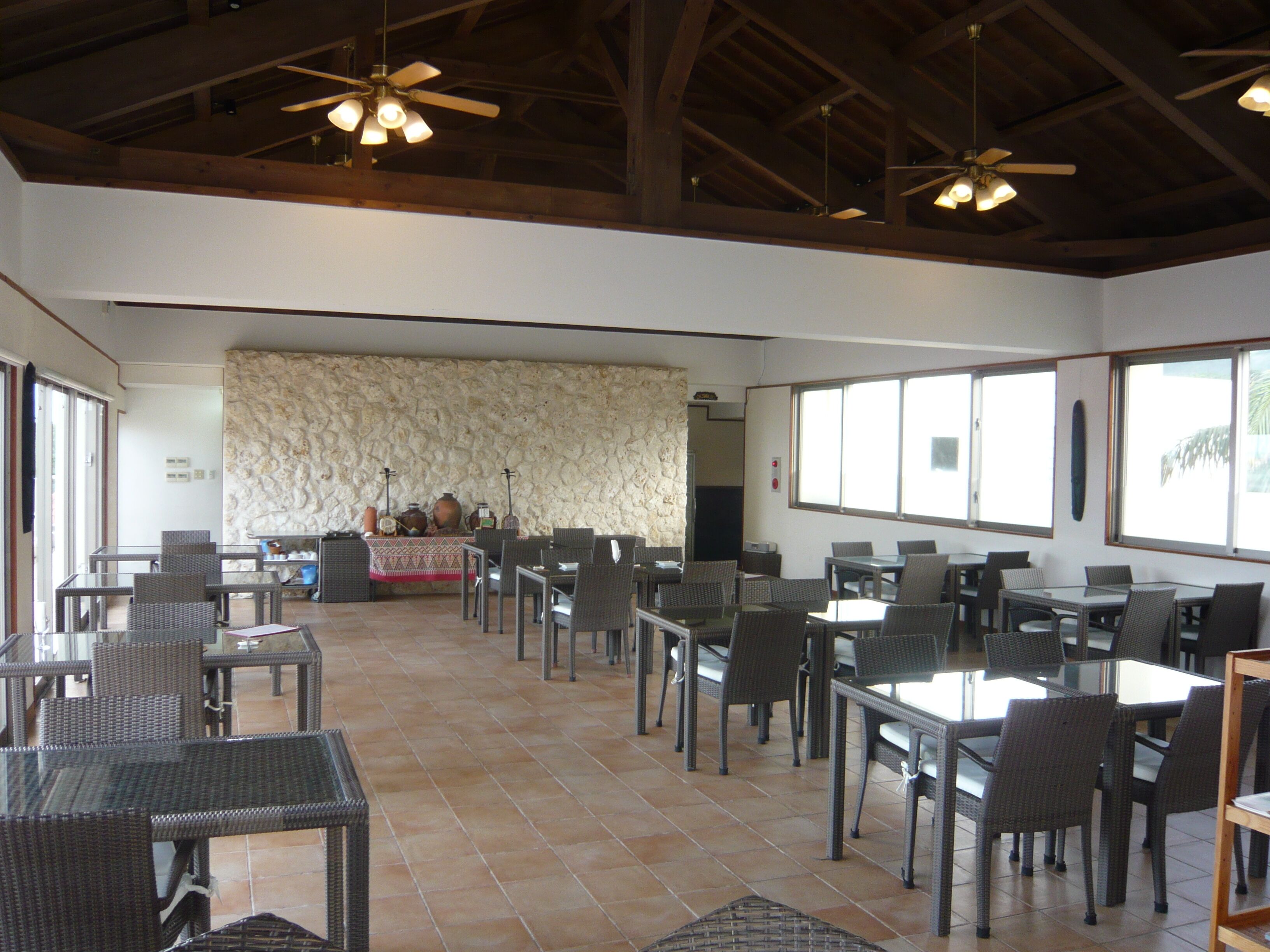 Un comedor interior amplio con varias mesas y sillas, una pared de piedra y ventanas grandes.