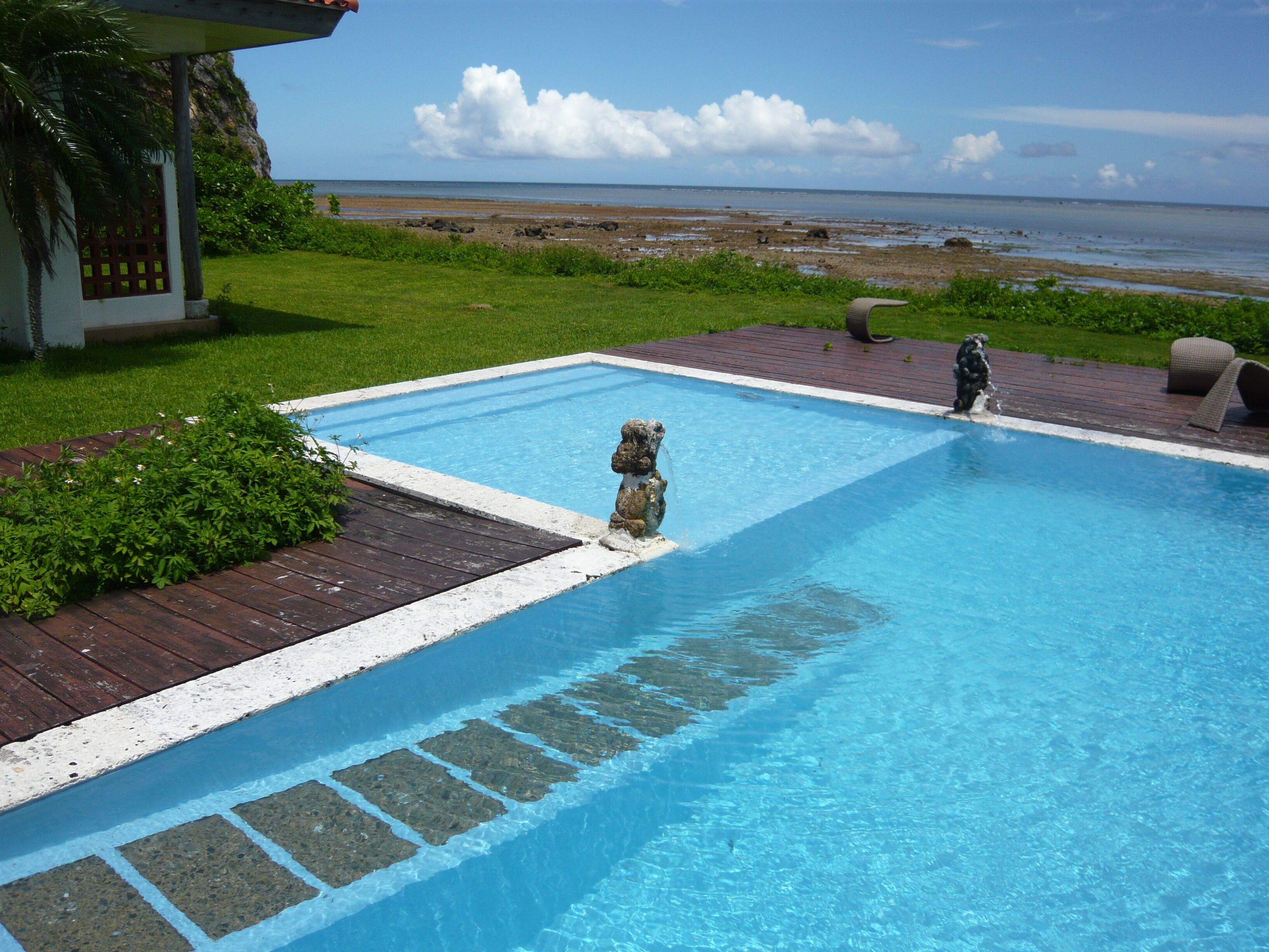 A swimming pool with a stone statue, a wooden deck, and a view of the beach.