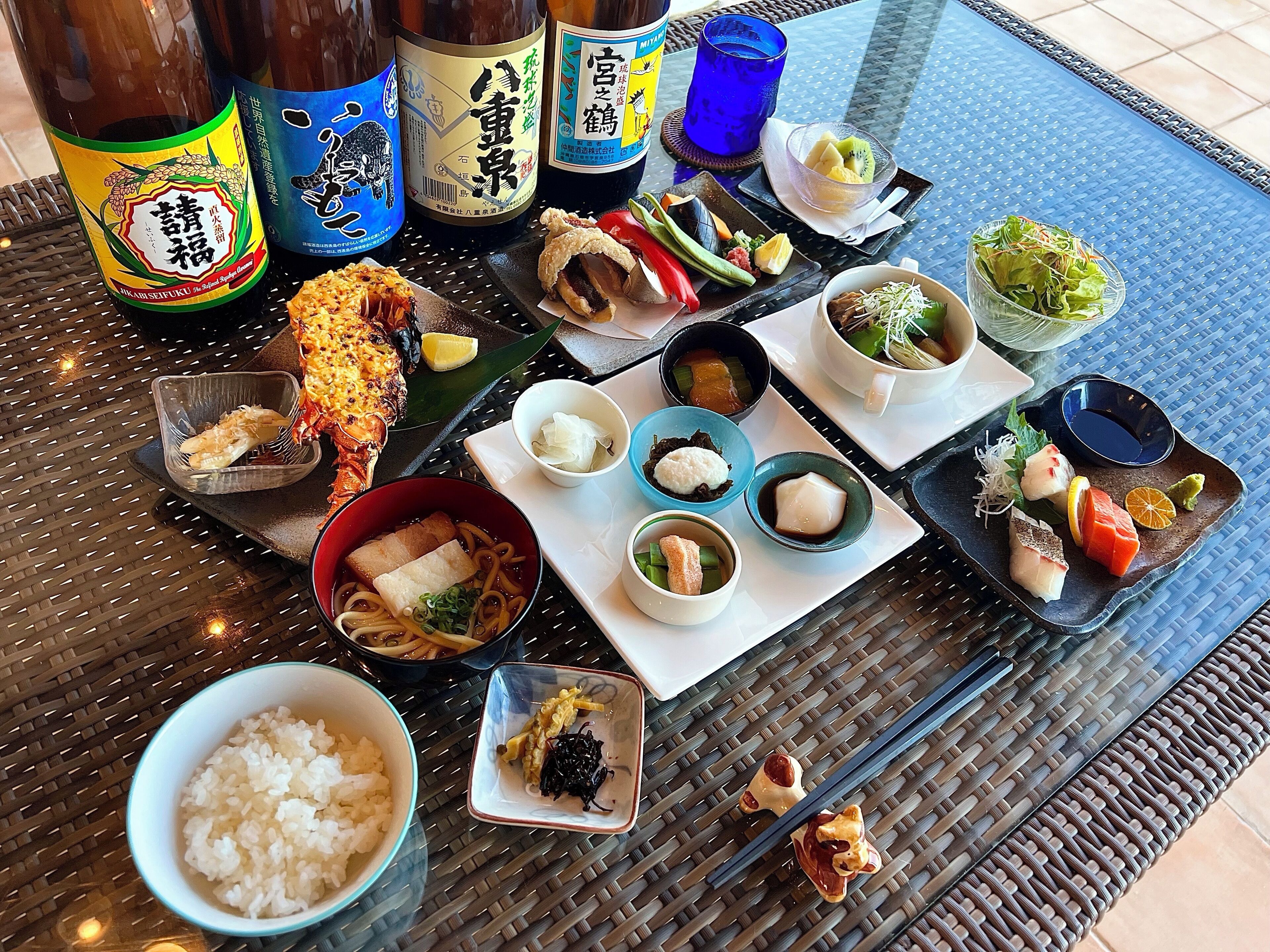 A table with various Japanese dishes, including sushi, rice, and a grilled fish, along with several bottles of beverages.