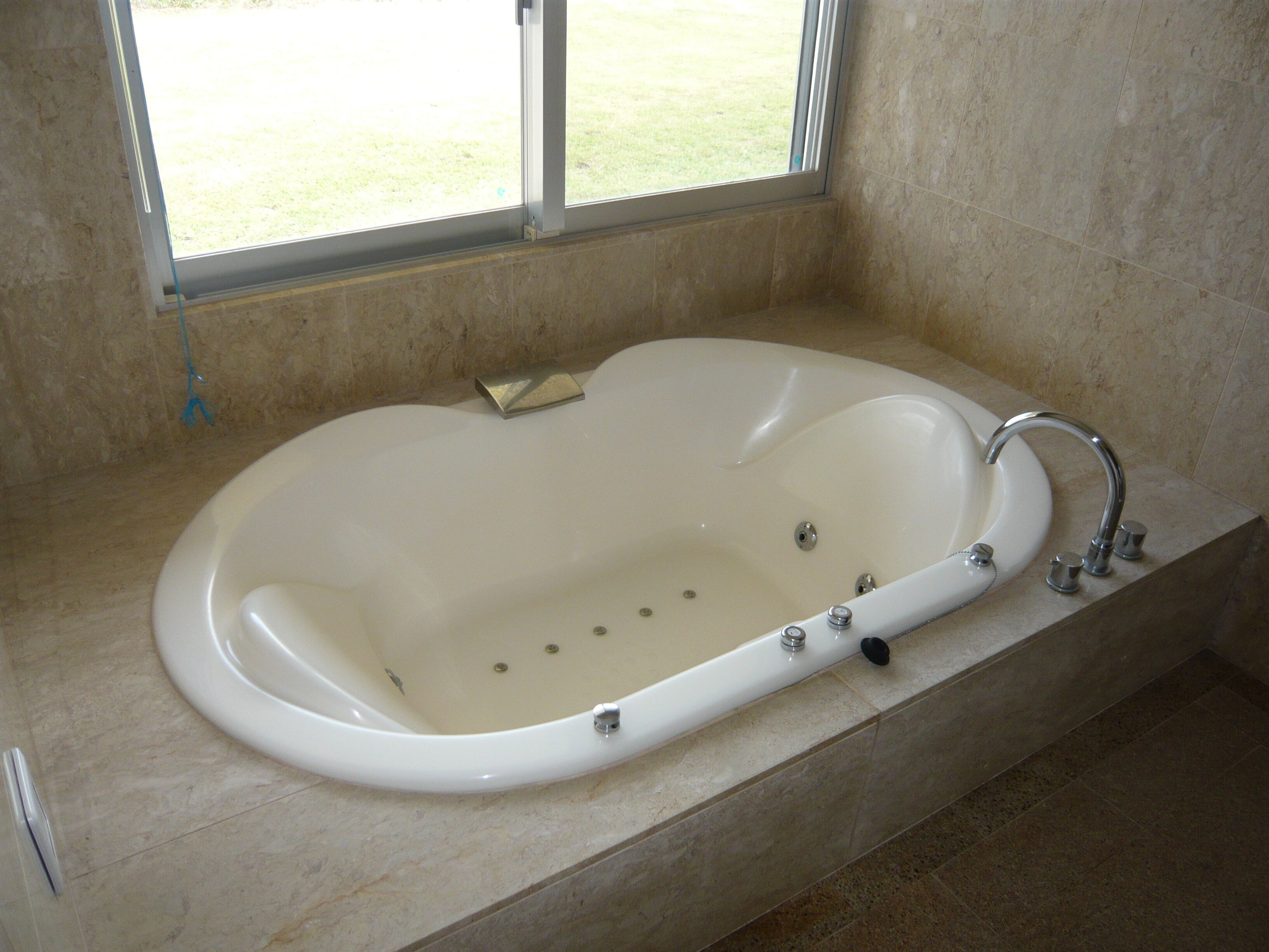 Une grande baignoire de forme ovale, dotée d’un seul robinet et d’une douchette à main, installée dans une salle de bain carrelée de carreaux beiges et comportant une fenêtre donnant sur un espace verdoyant.