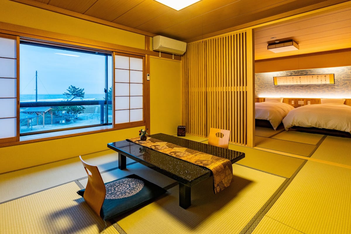 A traditional Japanese room with tatami flooring, a low table with a cushion, a futon bed, and a view of the sea through shoji screens.