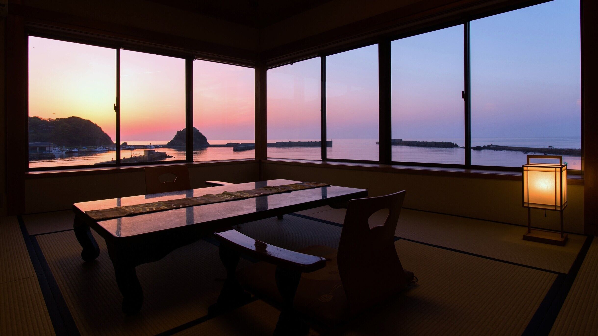 A room with a table and chairs, large windows with a view of the sea and a sunset, and a floor lamp.