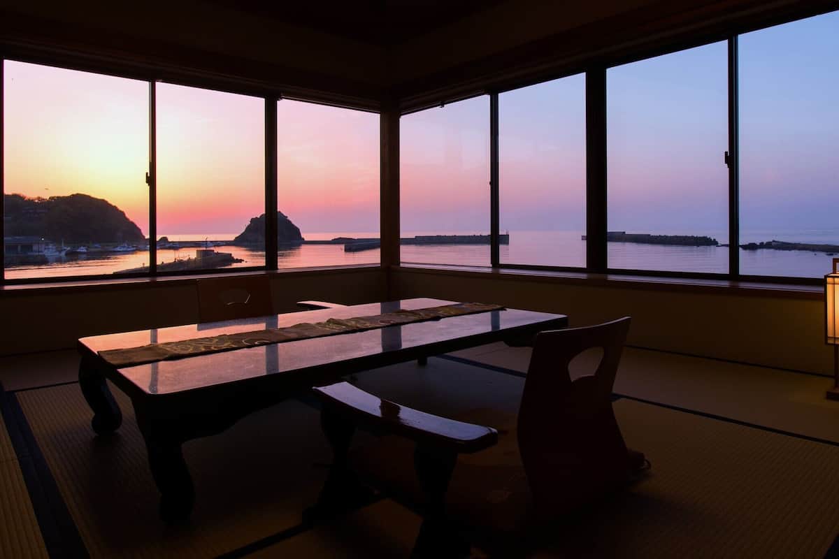 A room with a table and chairs, large windows with a view of the sea and a sunset, and a floor lamp.
