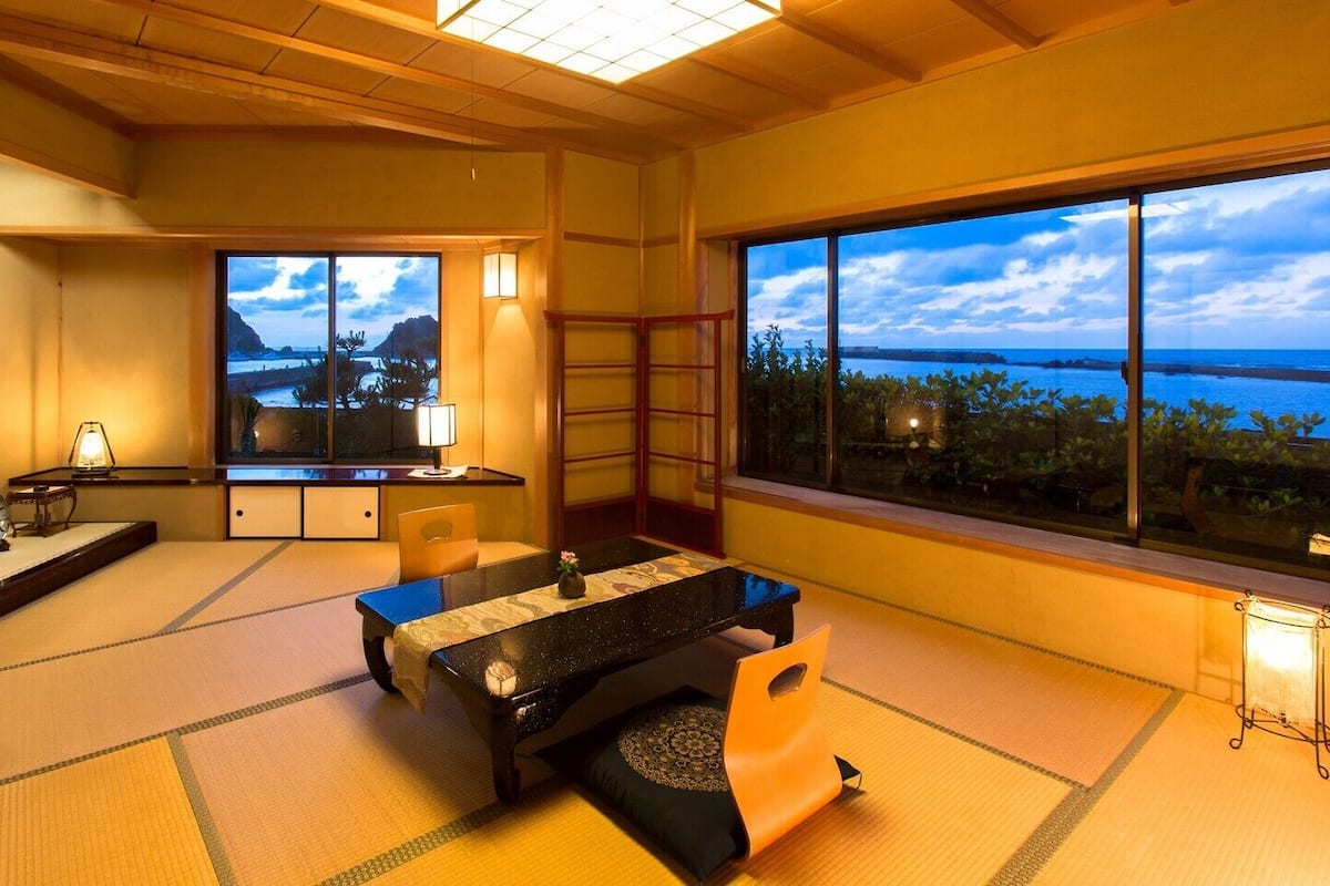 A traditional Japanese room with tatami flooring, a low table, a chair, and a view of the sea.