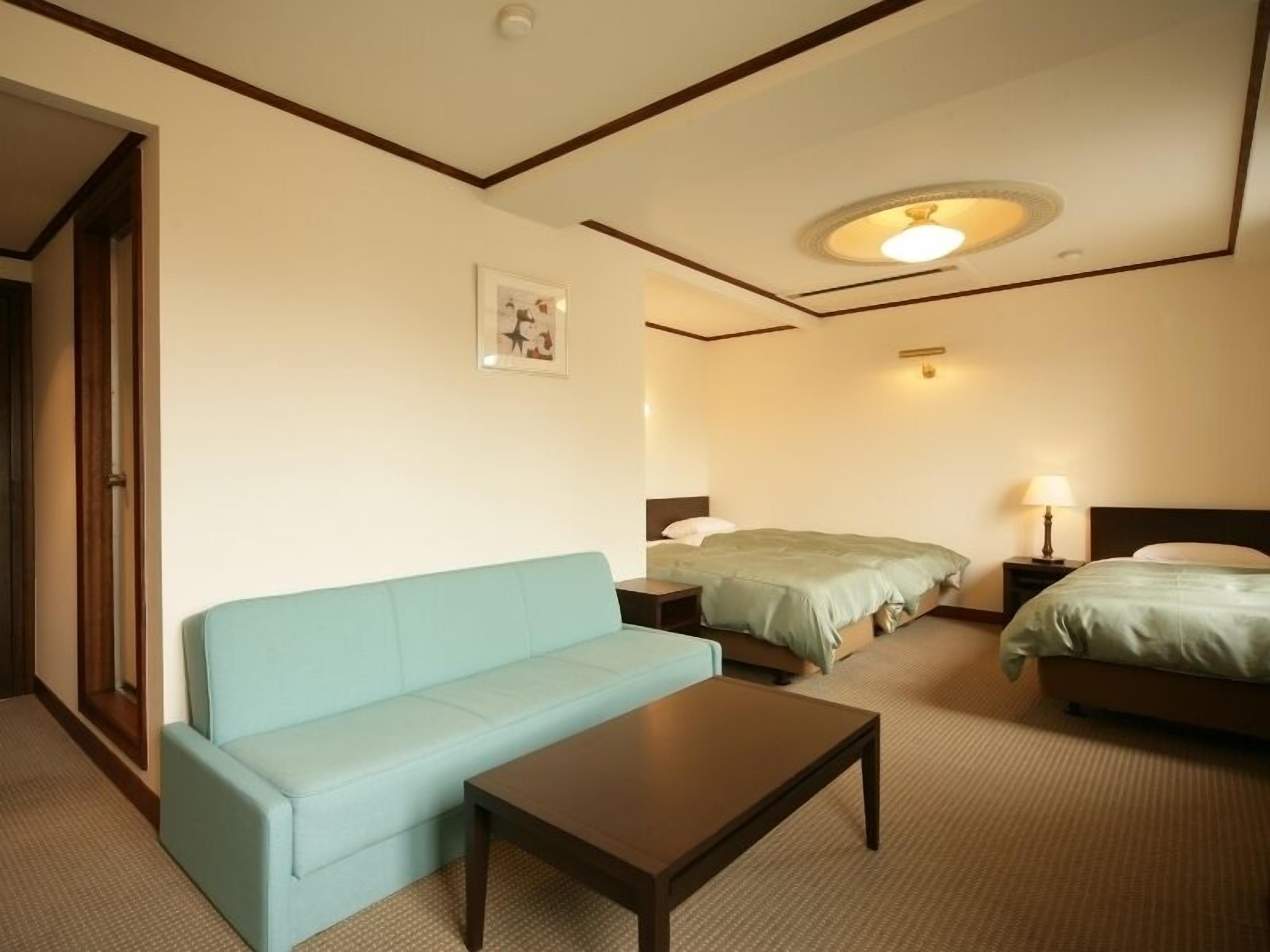 A hotel room with a light blue sofa, a wooden coffee table, two beds with green bedding, a nightstand with a lamp, and a ceiling light fixture.