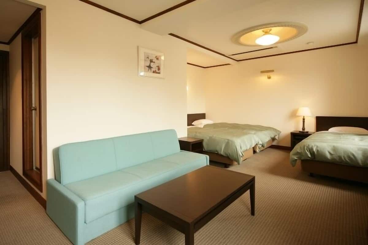 Habitación de hotel con un sofá azul claro, una mesa de centro de madera, dos camas con ropa de cama verde, una mesita de noche con lámpara y un candelabro de techo.