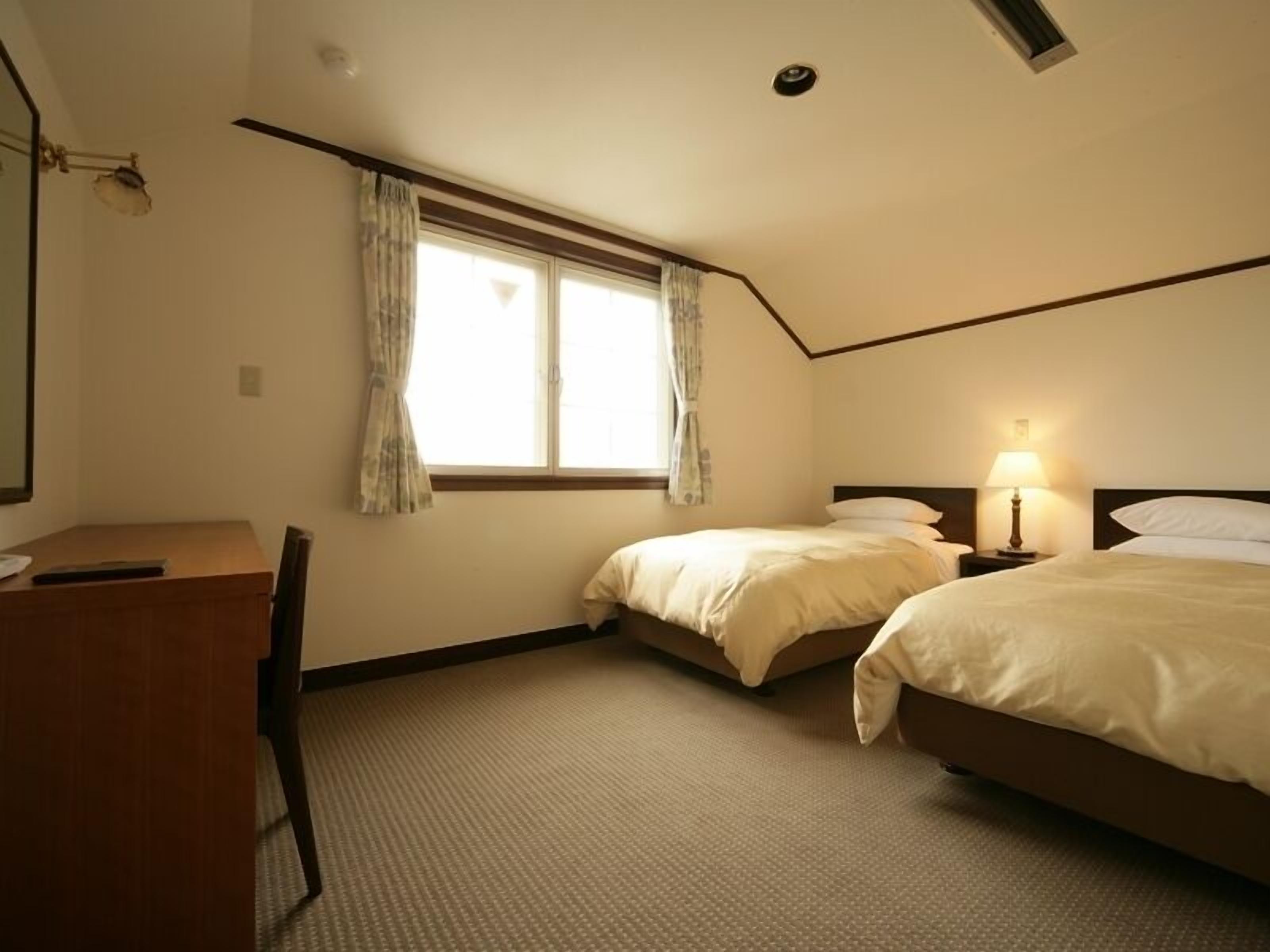 A hotel room with two beds, a desk, a lamp, and a window with curtains.